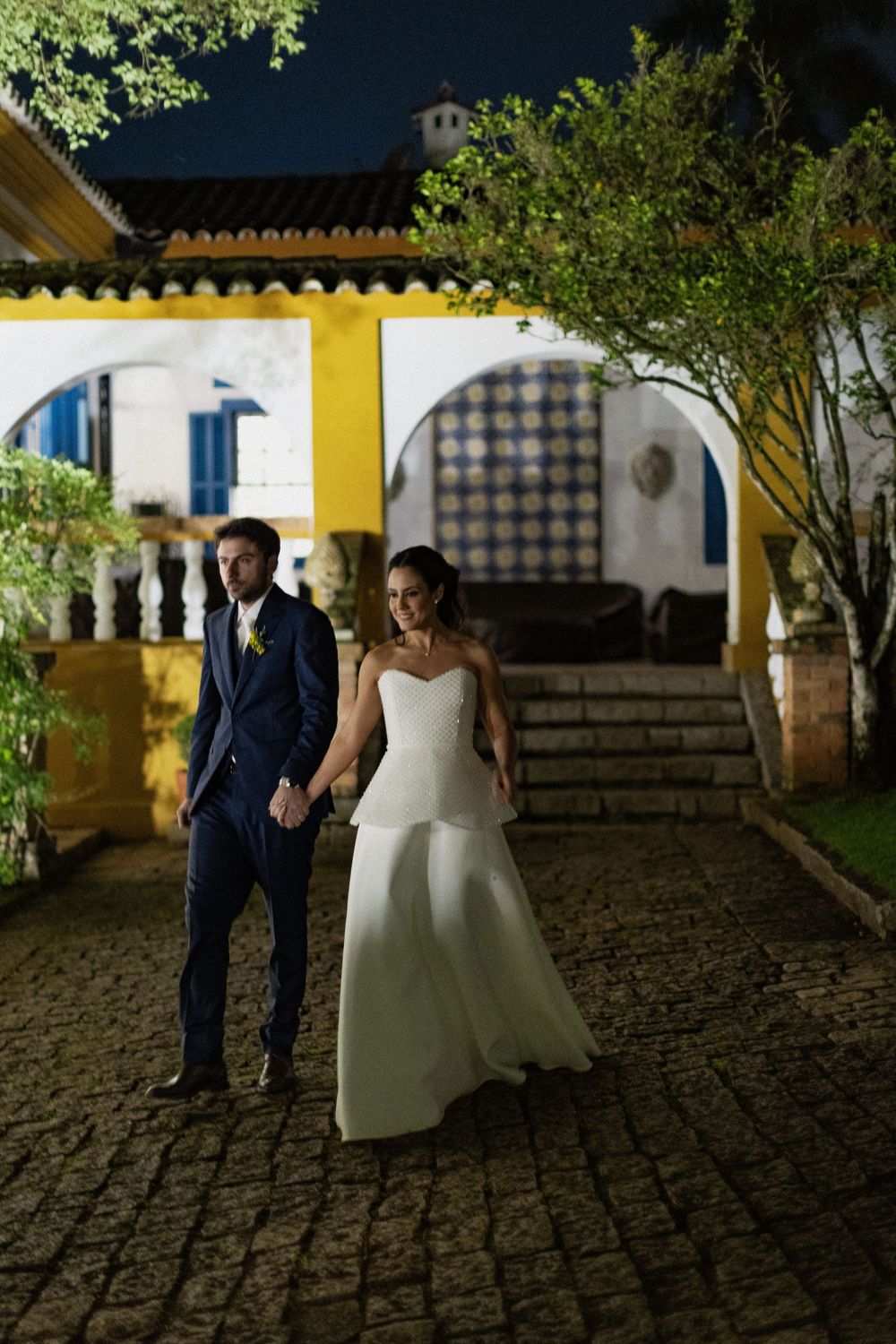 Bride in a strapless white gown and groom in a navy suit hold hands on a cobblestone path outside a yellow villa at night.