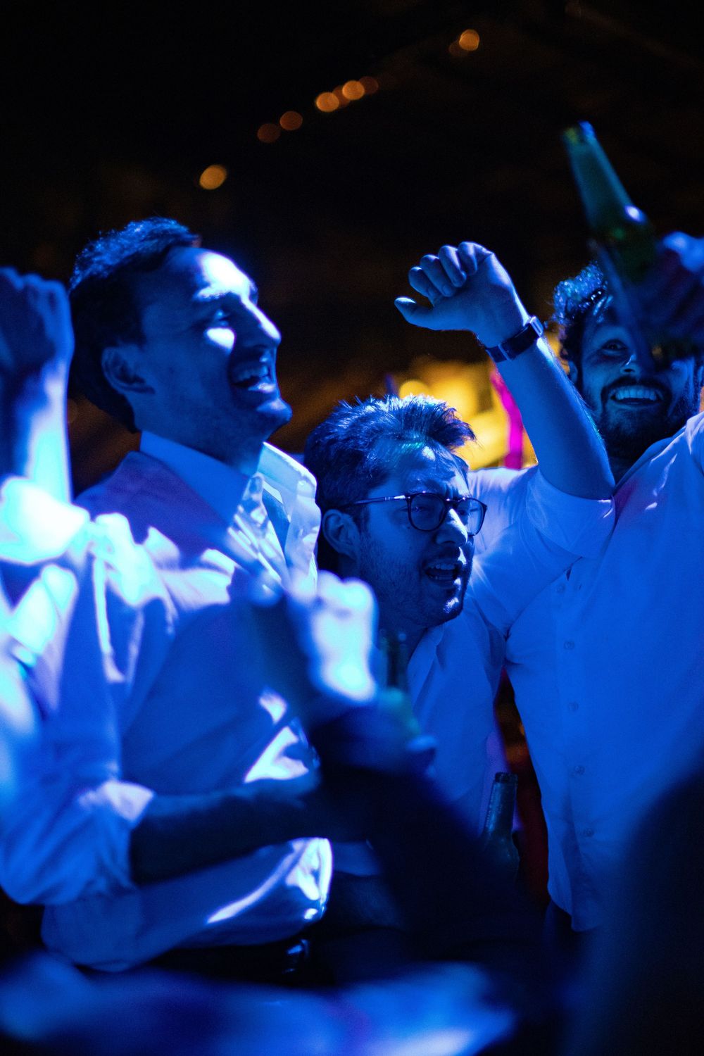 Group of men in white shirts dancing and celebrating at a lively nighttime party under blue lighting, raising their fists and smiling.