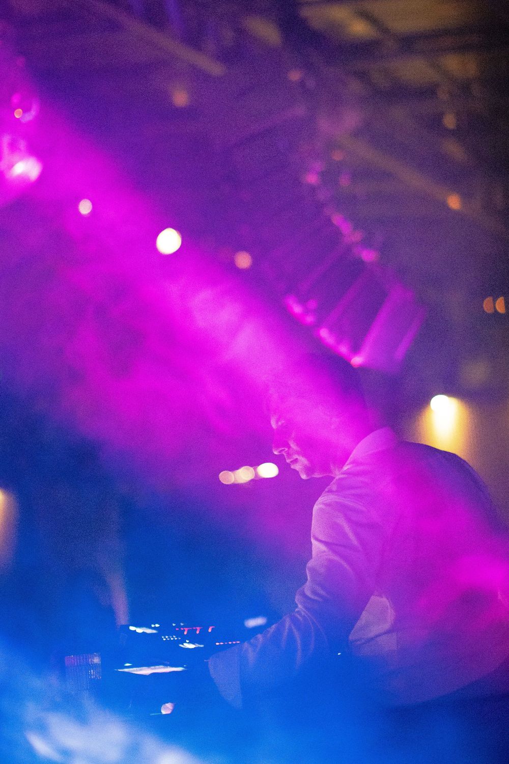 DJ performing at a nightclub behind the mixing console, surrounded by haze and vibrant purple and blue stage lighting.