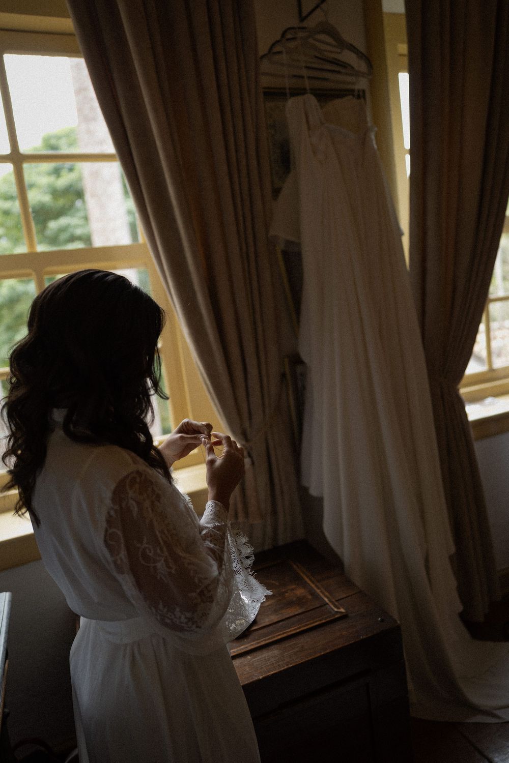 Bride in a white lace robe stands by a sunlit window, holding jewelry while her wedding dress hangs nearby.