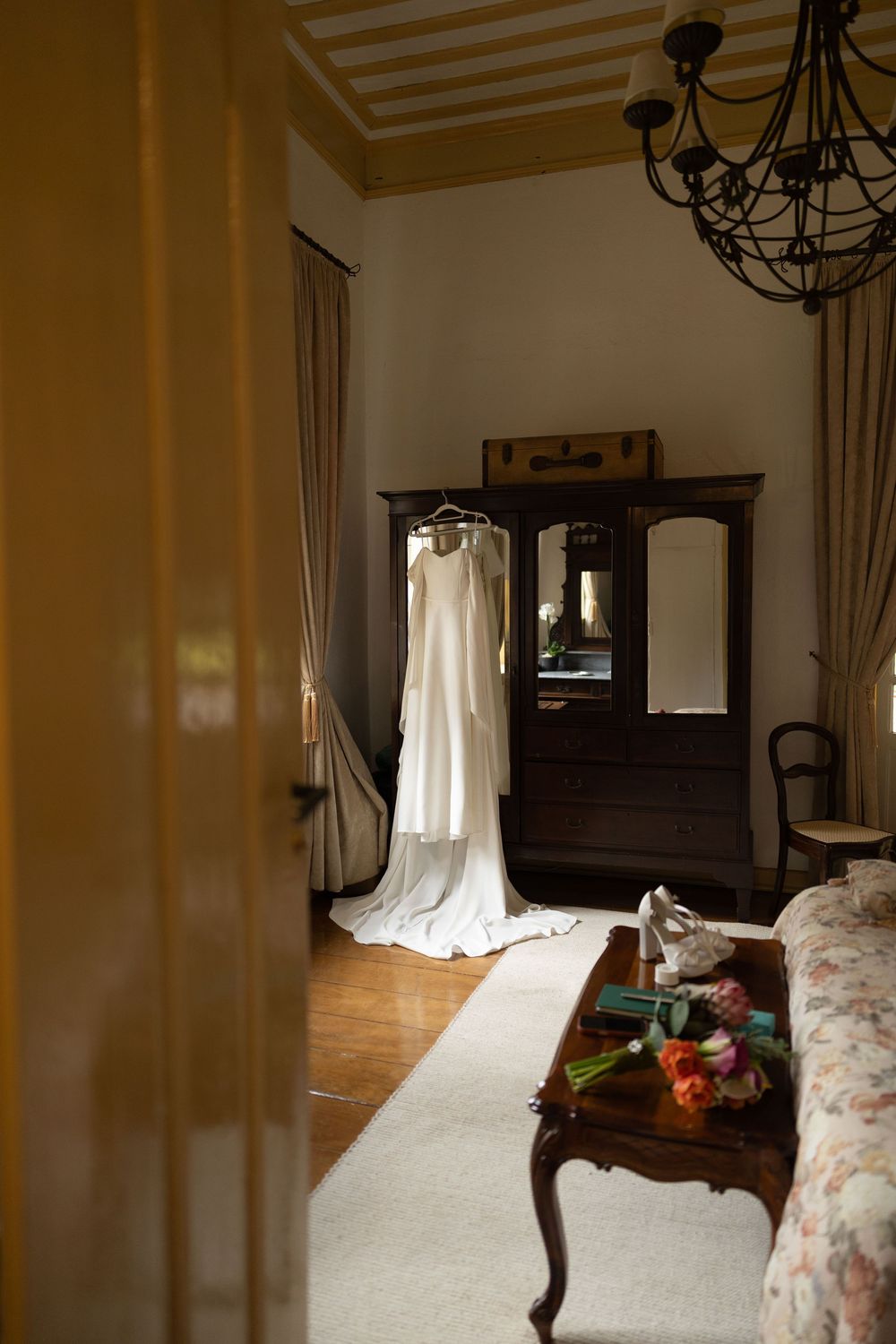 White wedding dress hanging on a wooden armoire with mirrors in an elegant bedroom with a chandelier, bridal bouquet and shoes on a table.