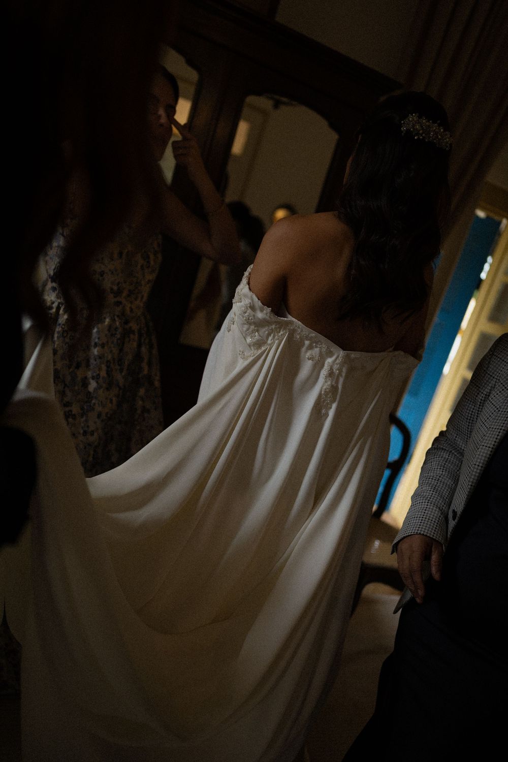 Bride in an off-the-shoulder white wedding gown with a flowing train and a hair accessory, seen from behind in a dimly lit room with guests.