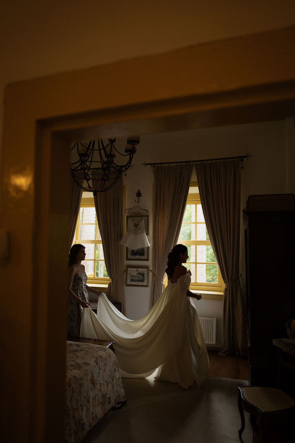 Bride in a flowing white wedding gown with a long train stands by a sunlit window in an elegant bedroom while another woman looks on.