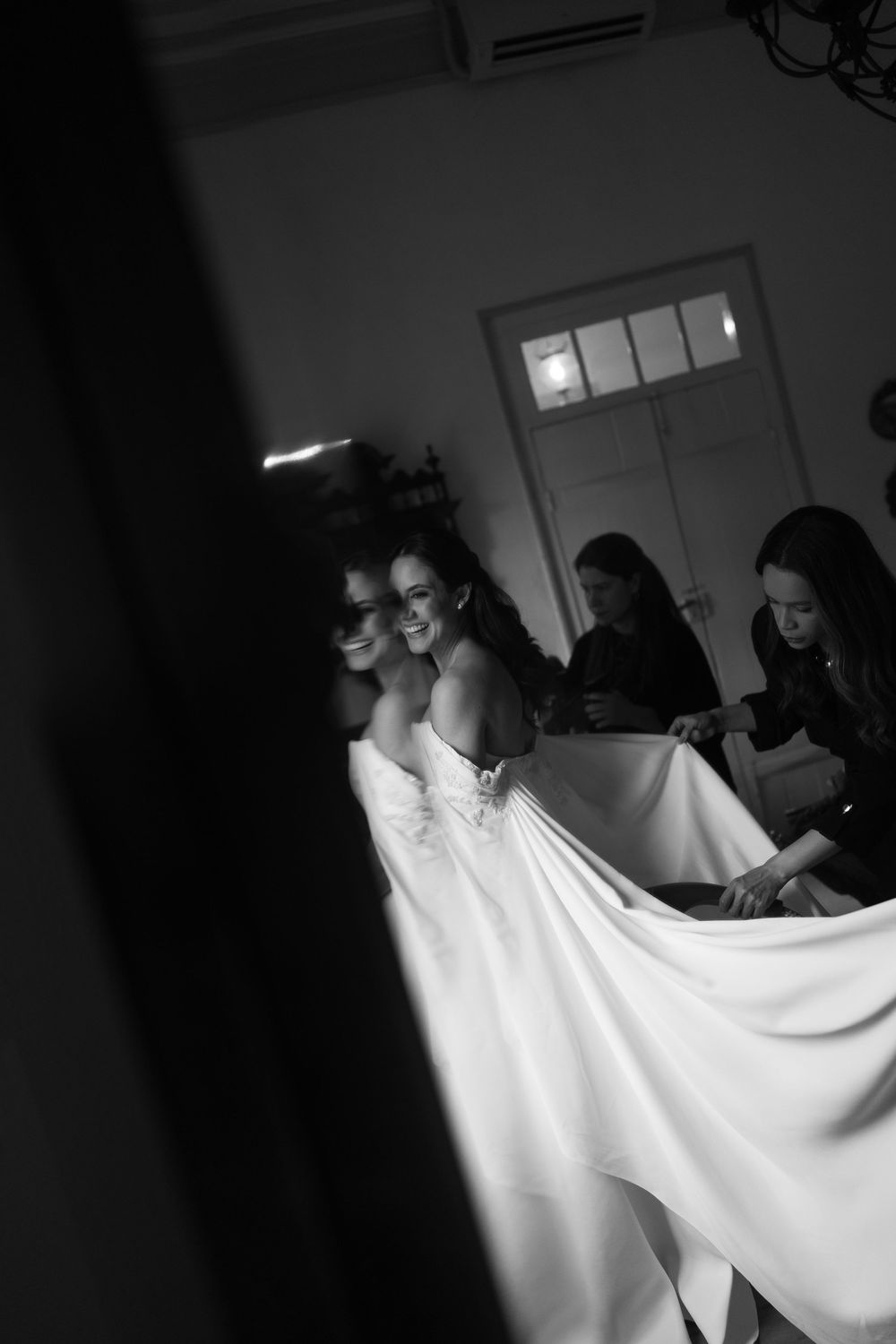 Black and white photo of a smiling bride in a strapless white wedding gown while attendants arrange her flowing train in a softly lit room.