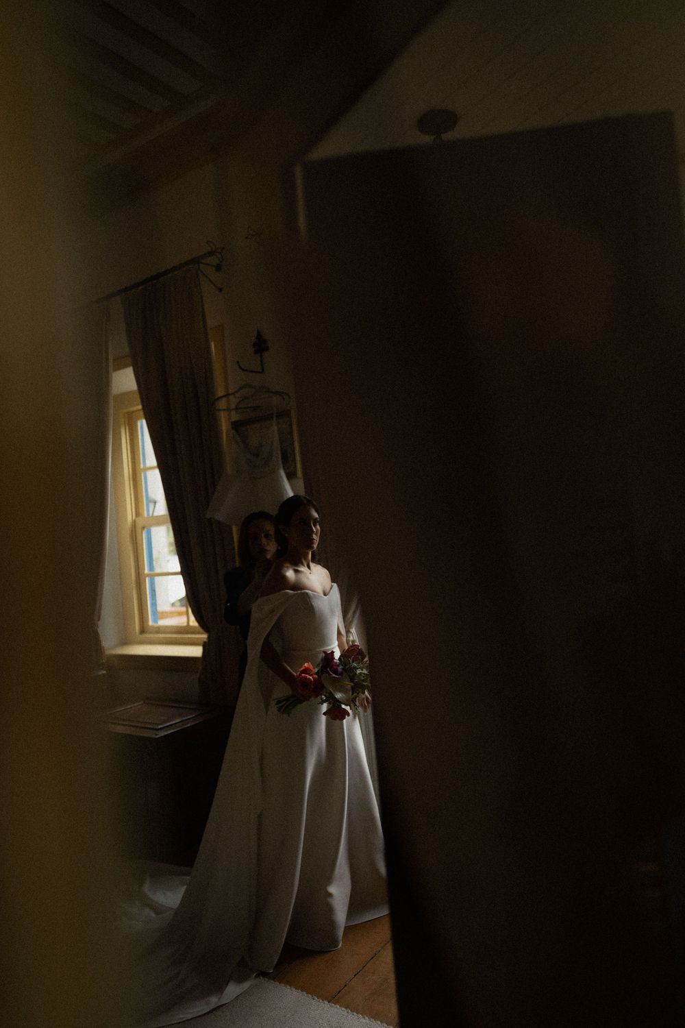 Bride in a flowing white gown holding a colorful bouquet stands by a sunlit window in a softly shadowed, rustic room with wooden floors.
