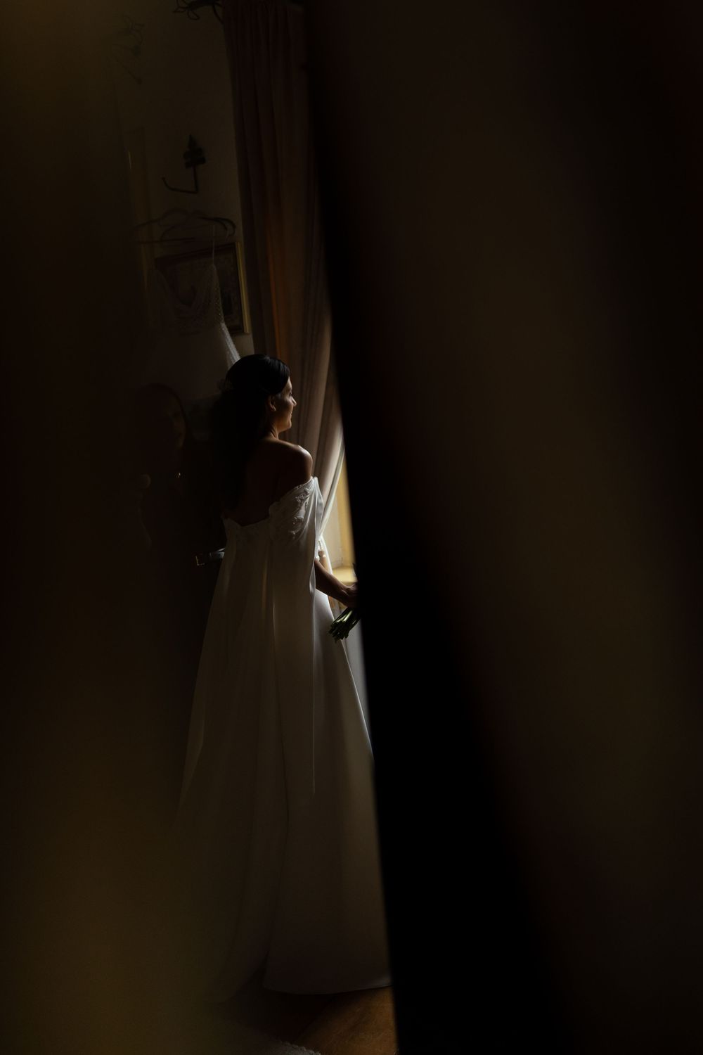 Bride in a white off-the-shoulder gown stands by a sunlit window, framed by dark curtains in a moody, dramatic indoor wedding portrait.