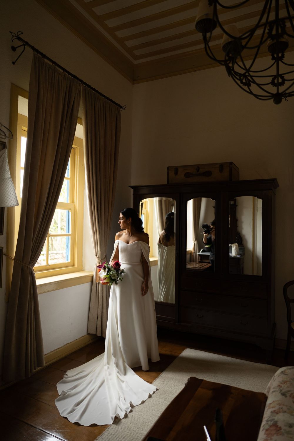 Bride in an off-shoulder white gown with a long train holds a bouquet beside a sunlit window in an elegant room with a mirrored wardrobe.