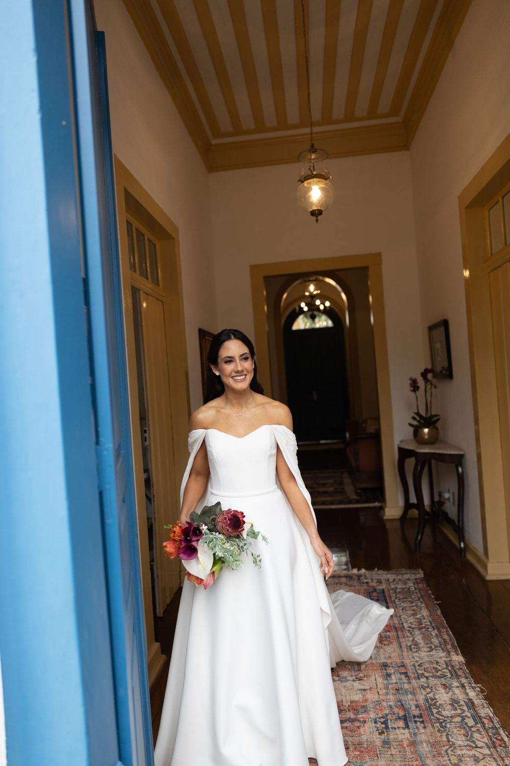 Smiling bride in an off-the-shoulder white wedding gown holding a colorful bouquet while walking down a bright hallway with a vintage rug.