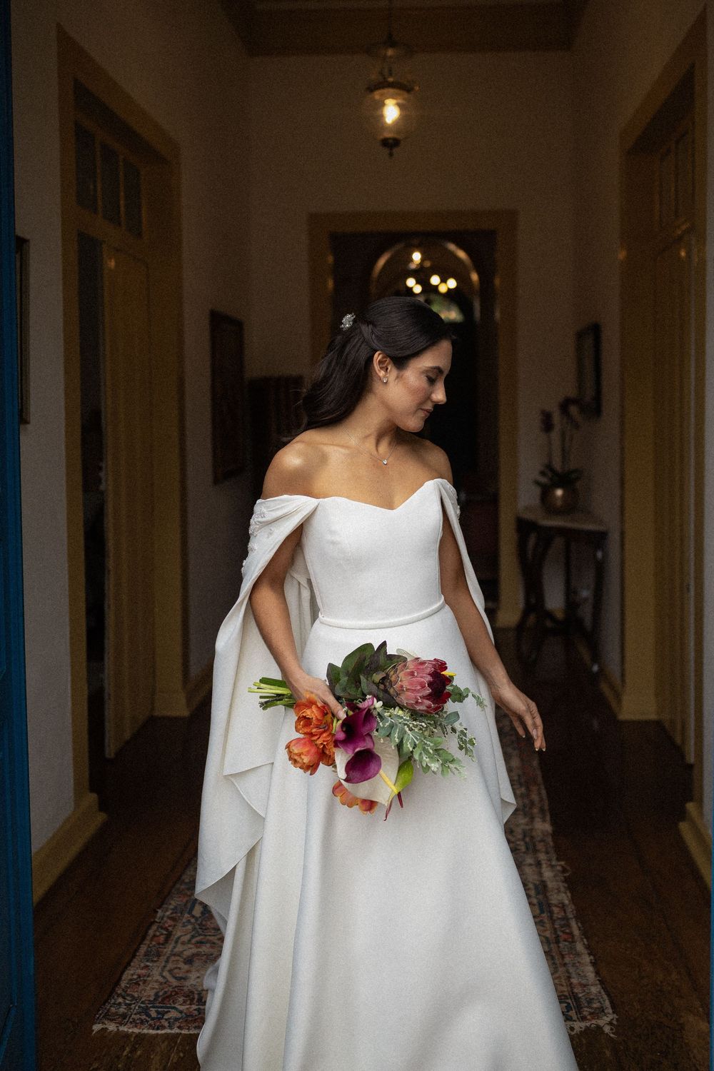 Bride in an off-the-shoulder white wedding gown with cape sleeves holds a colorful bouquet while standing in a warm, elegant hallway.