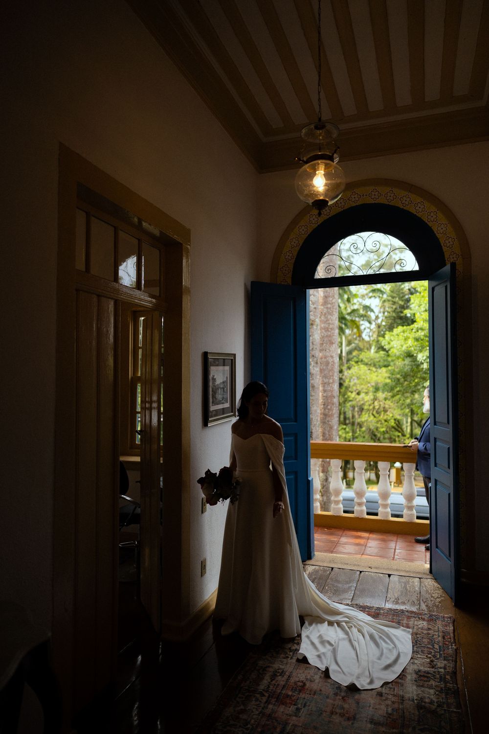Bride in an off-the-shoulder wedding gown with a long train stands holding a bouquet beside an open blue door overlooking a sunlit garden.
