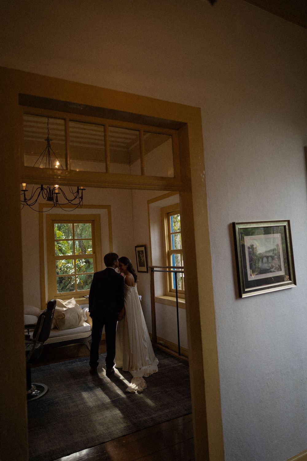 Bride and groom embrace in a sunlit room with a chandelier and tall windows, framed by a wooden doorway in a romantic wedding portrait.