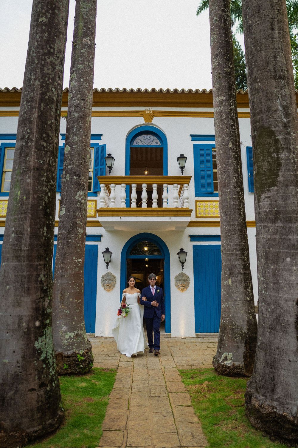 Bride in a white gown and groom in a navy suit walk arm in arm down a stone path between tall palm trees toward a white colonial building.