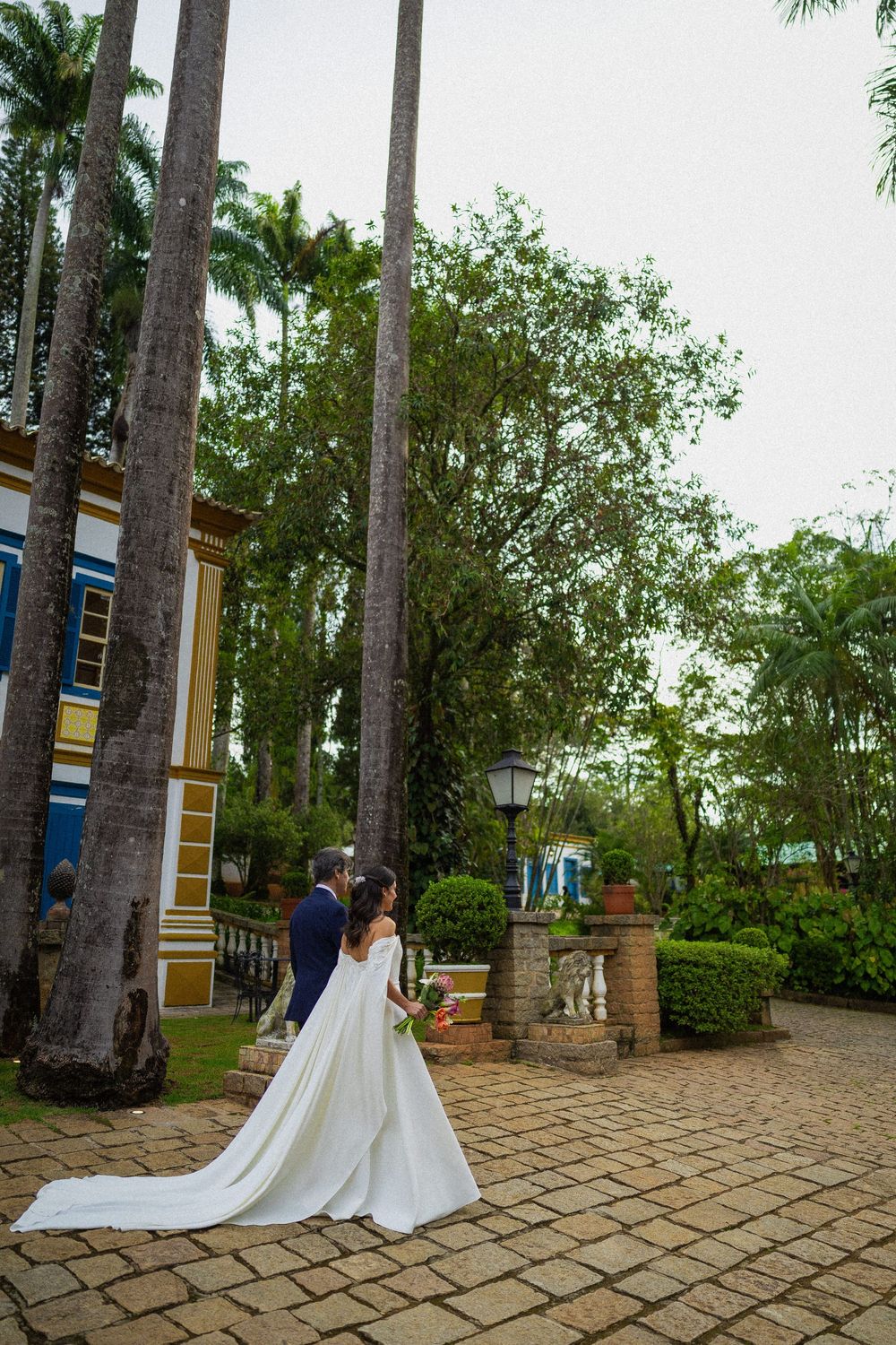 Bride in a flowing white gown and groom in a navy suit walk a cobblestone path lined with tall palm trees at a colonial-style estate.