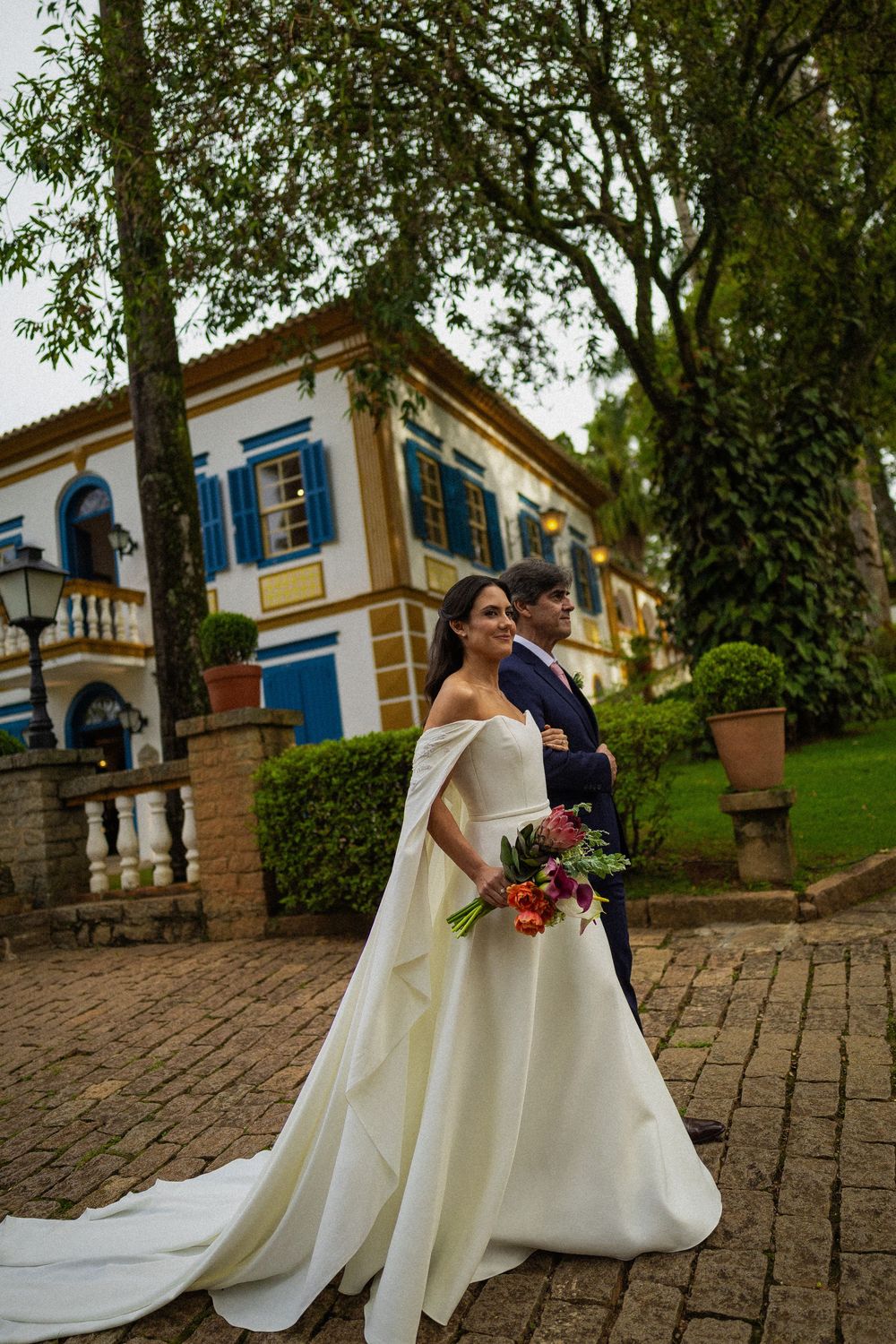 Bride in an off-shoulder white gown with a cape holding a colorful bouquet beside her groom on a brick path near a colonial-style building.