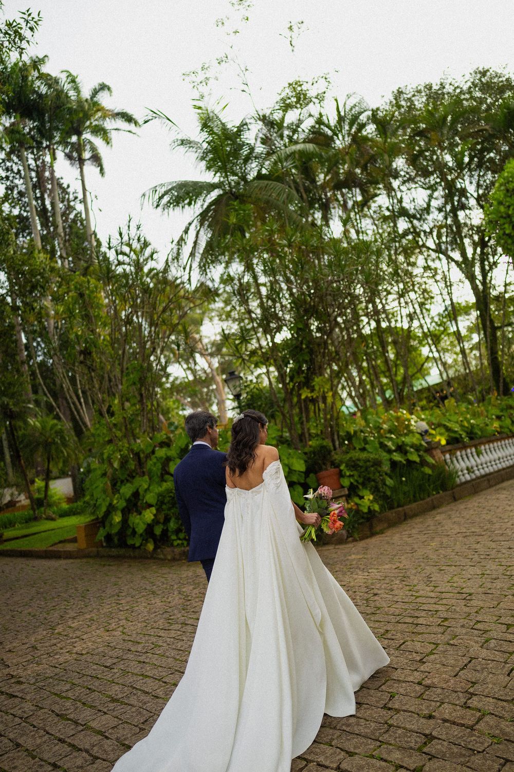 Bride in a flowing white gown and groom in a dark suit walk away along a brick path lined with lush palm trees and tropical greenery.