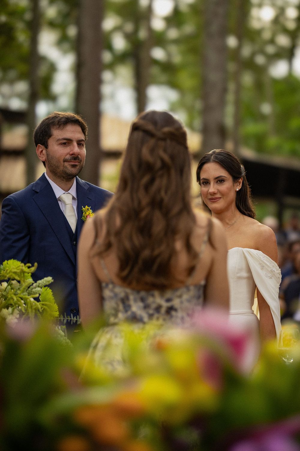 Groom in a navy suit and bride in a white gown smile at an outdoor forest wedding ceremony as a bridesmaid in a floral dress faces them.