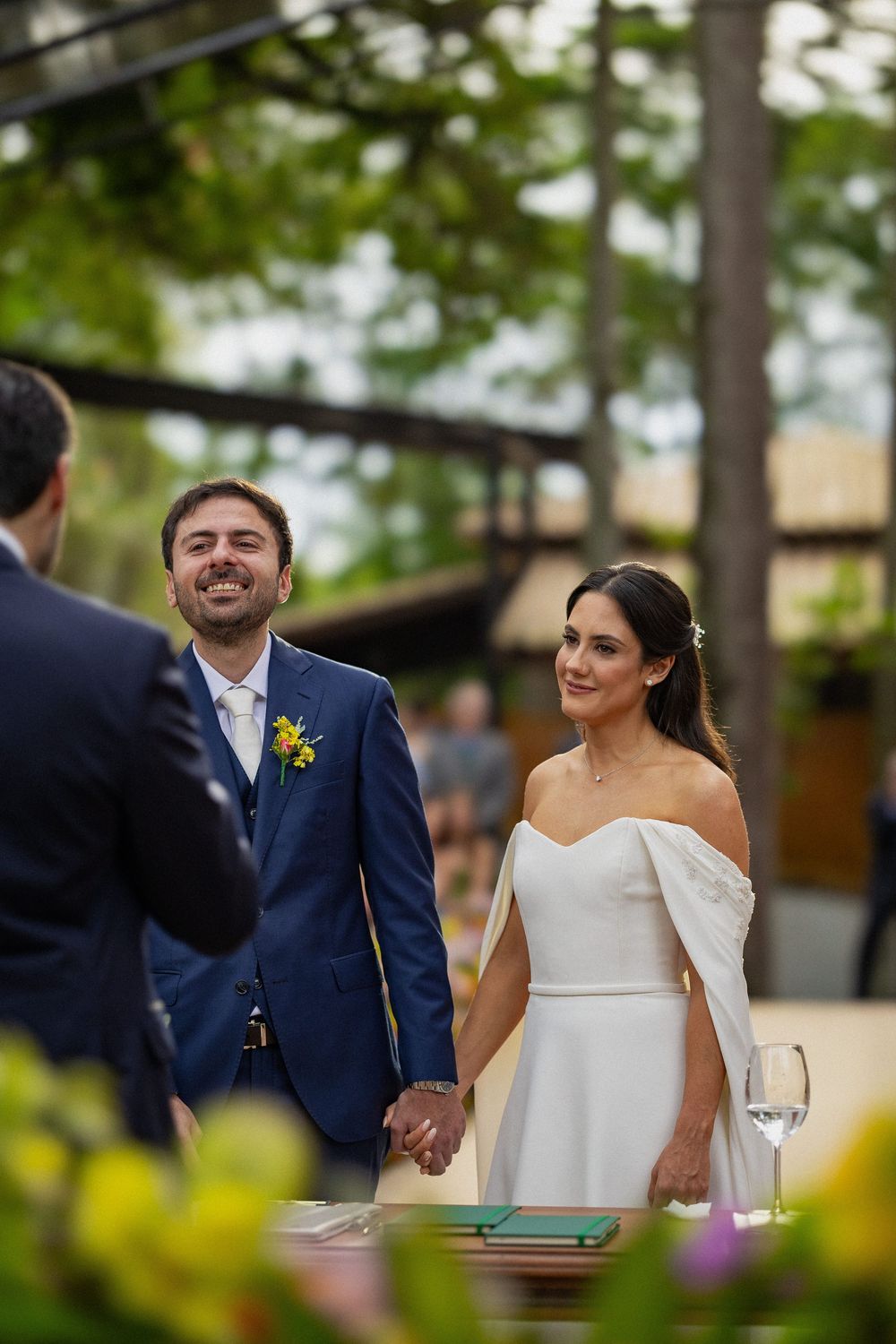 Smiling groom in a navy suit holds hands with the bride in a white gown during an outdoor wedding ceremony surrounded by trees.
