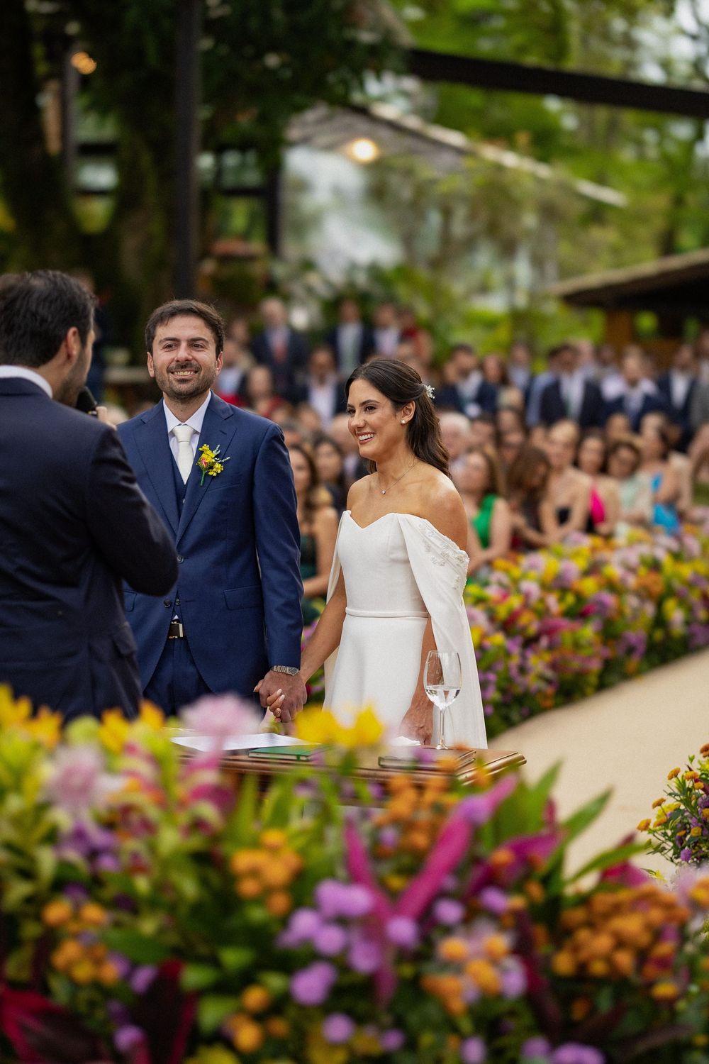 Smiling bride in an off-shoulder white gown holds hands with the groom during an outdoor wedding ceremony surrounded by colorful flowers and