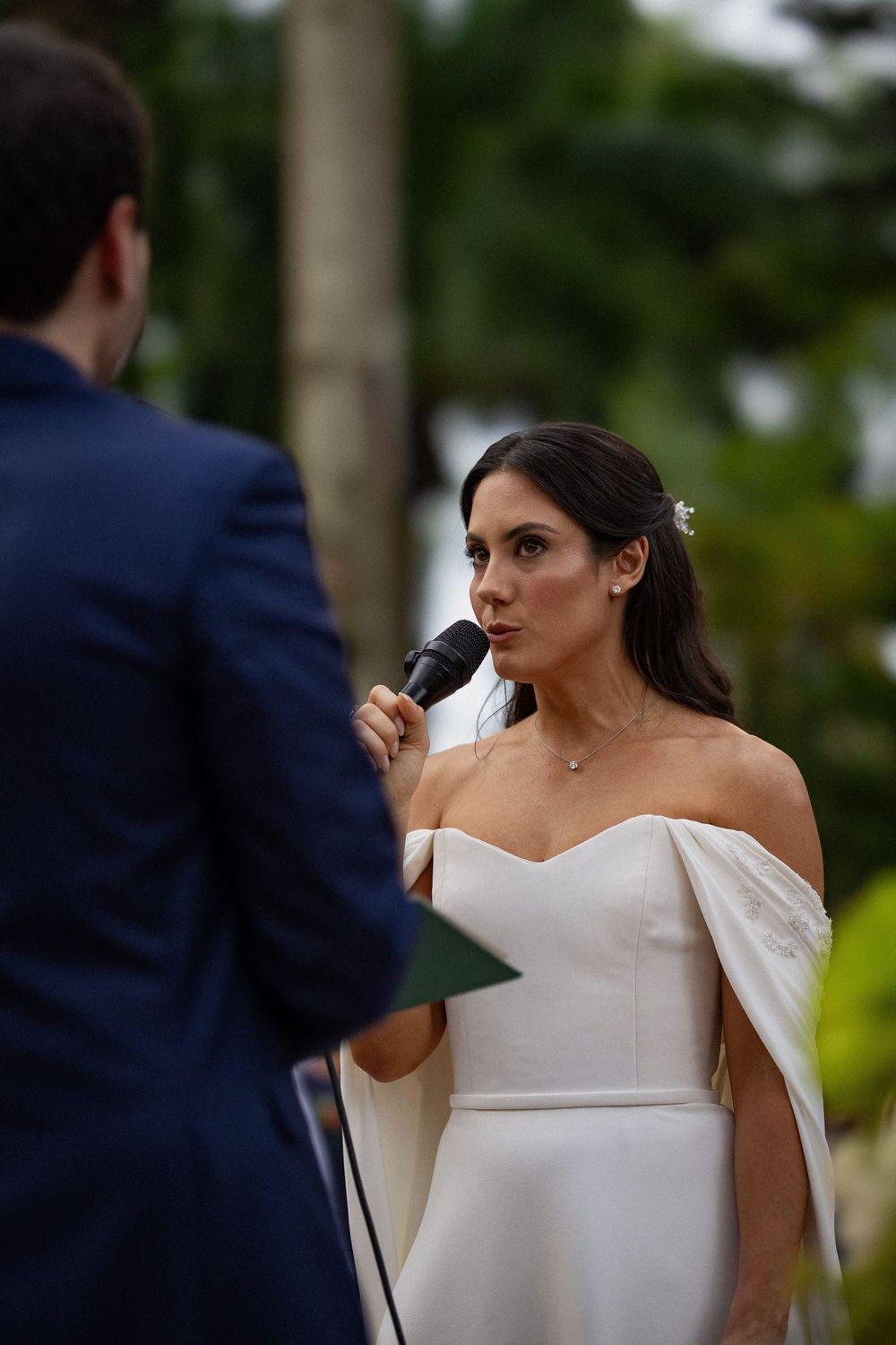 Bride in an off-the-shoulder white wedding gown speaks into a microphone during an outdoor ceremony, facing a groom in a navy suit.