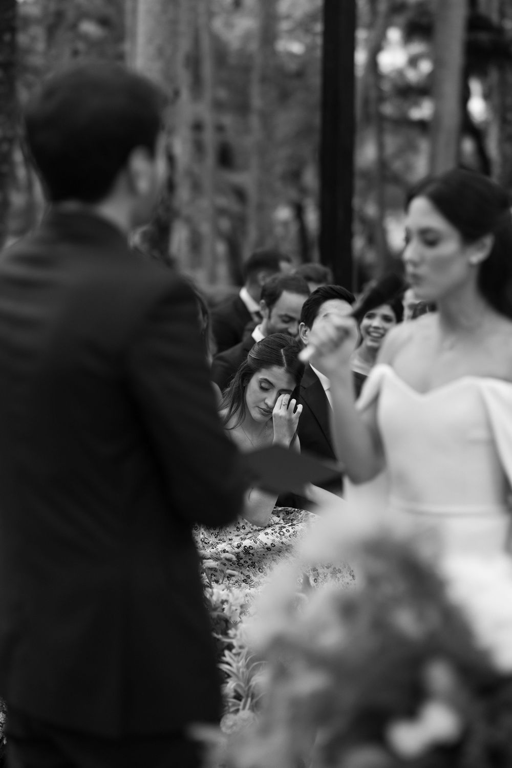Black and white wedding ceremony photo of a bride in an off-shoulder gown as emotional guests, one wiping tears, watch from the background.
