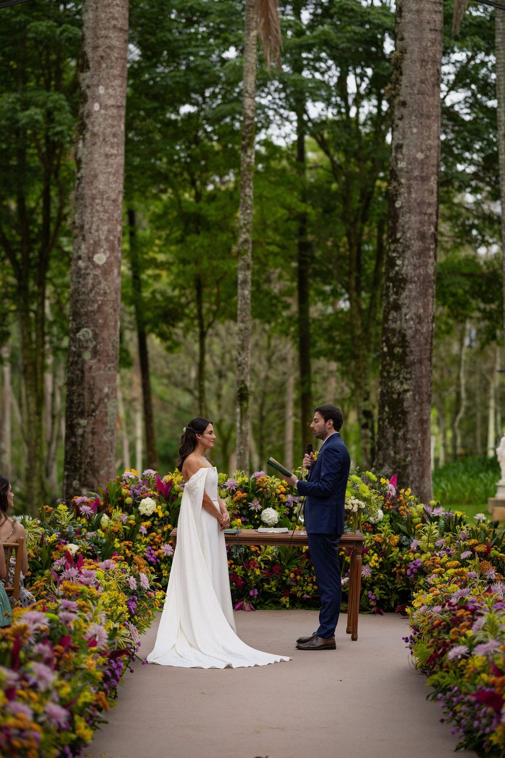 Bride in a white gown and groom in a navy suit exchange vows at a garden altar surrounded by colorful flowers and tall forest trees.
