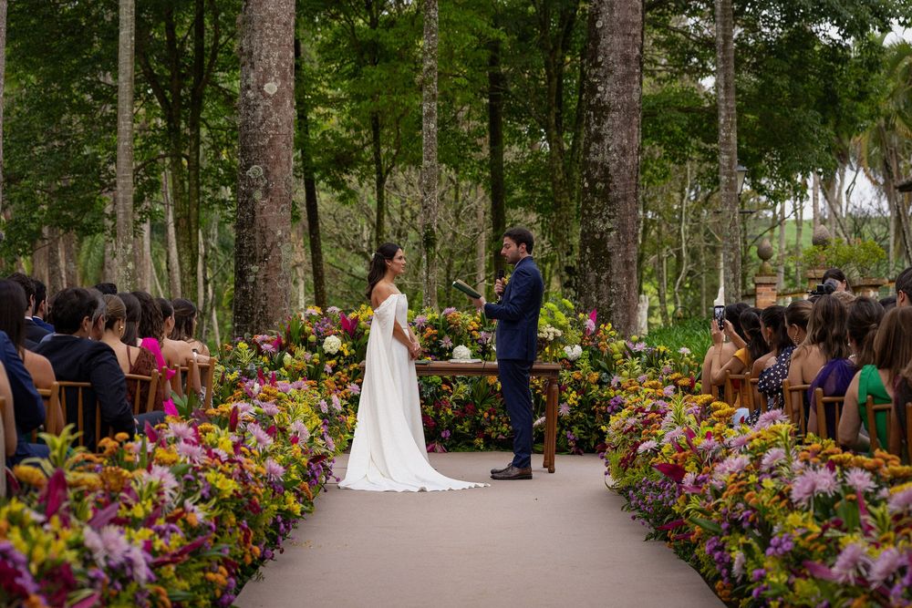 Bride and groom exchange vows at an outdoor forest wedding ceremony surrounded by colorful flower arrangements and seated guests.