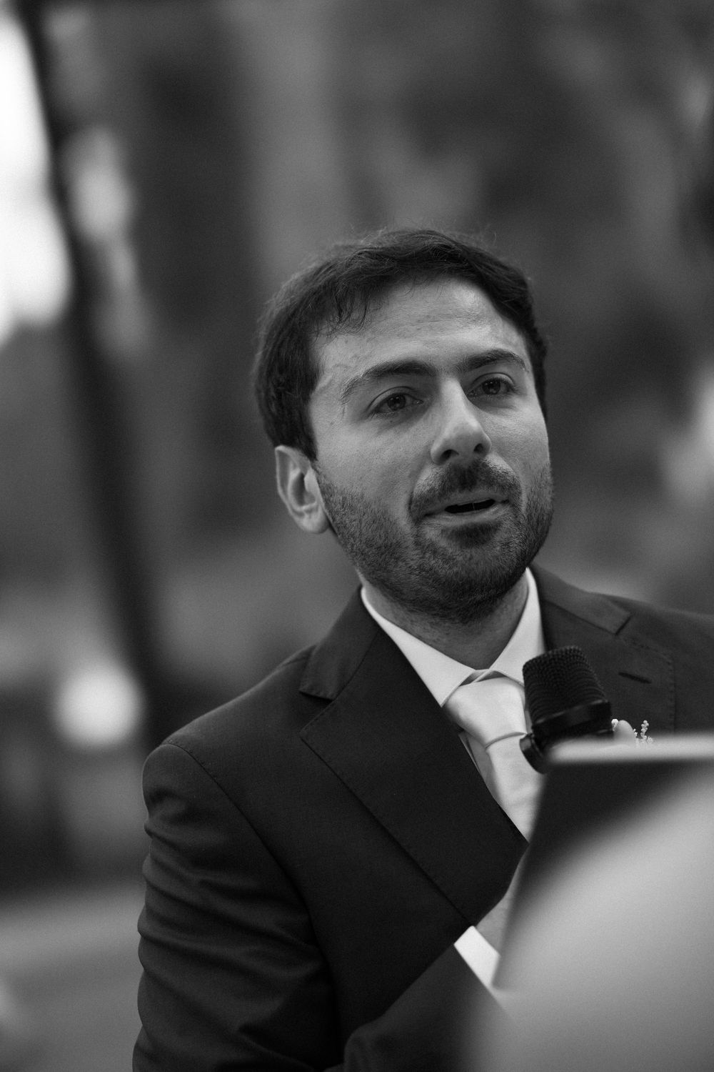 Black and white portrait of a bearded man in a suit and tie speaking into a microphone outdoors with a blurred background.