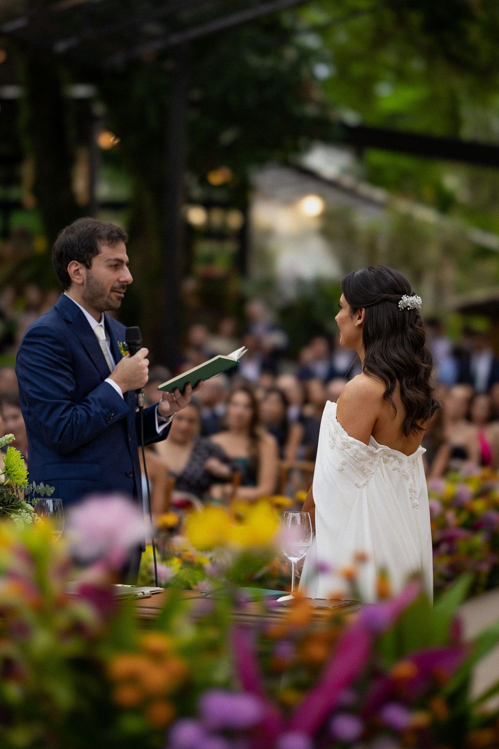 Groom in a navy suit reads from a book into a microphone as the bride in a white gown listens at an outdoor garden wedding ceremony.