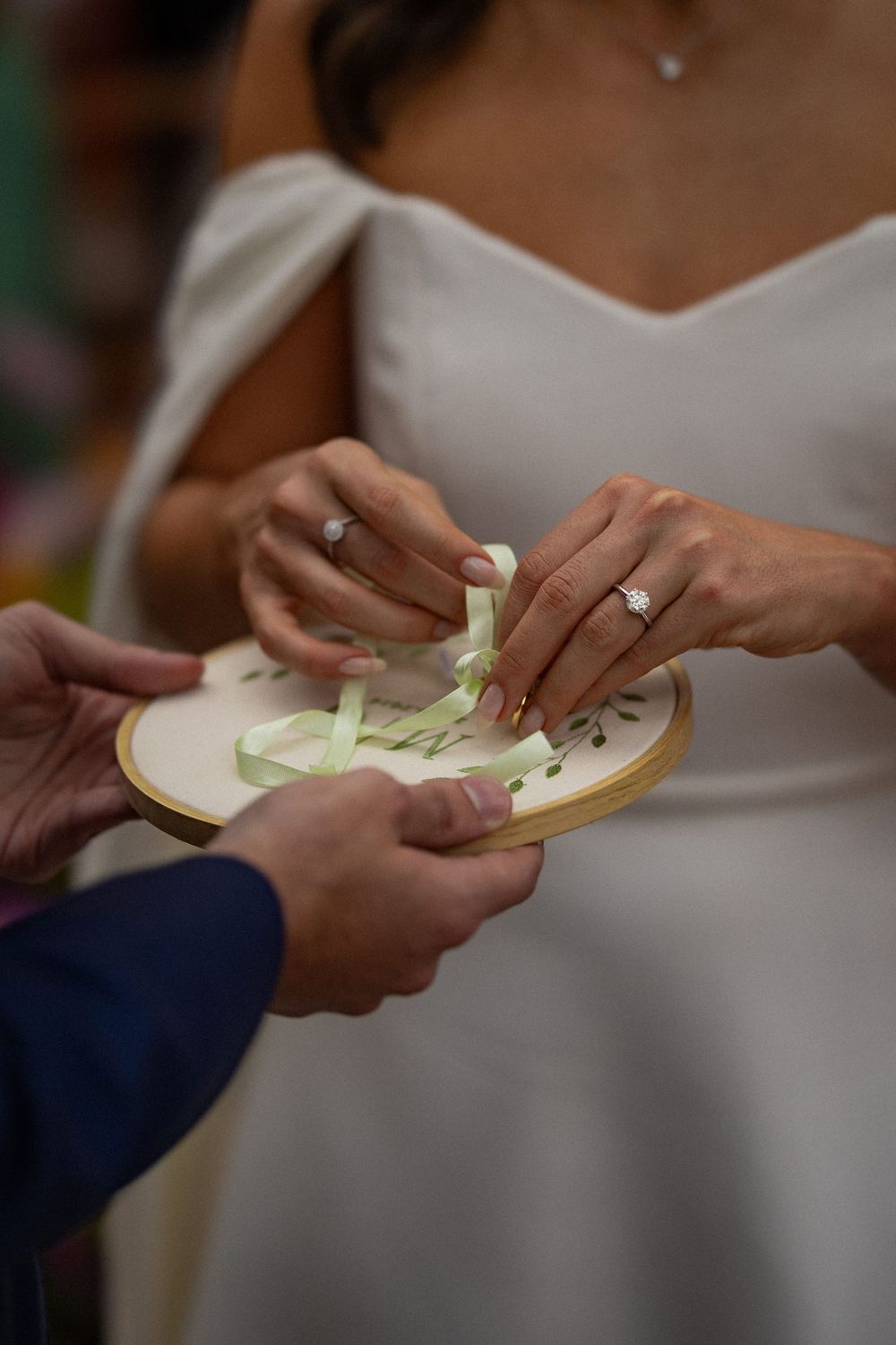 Bride in a white dress unties a green ribbon on a wooden embroidery hoop ring holder during a wedding ceremony as the groom holds it.