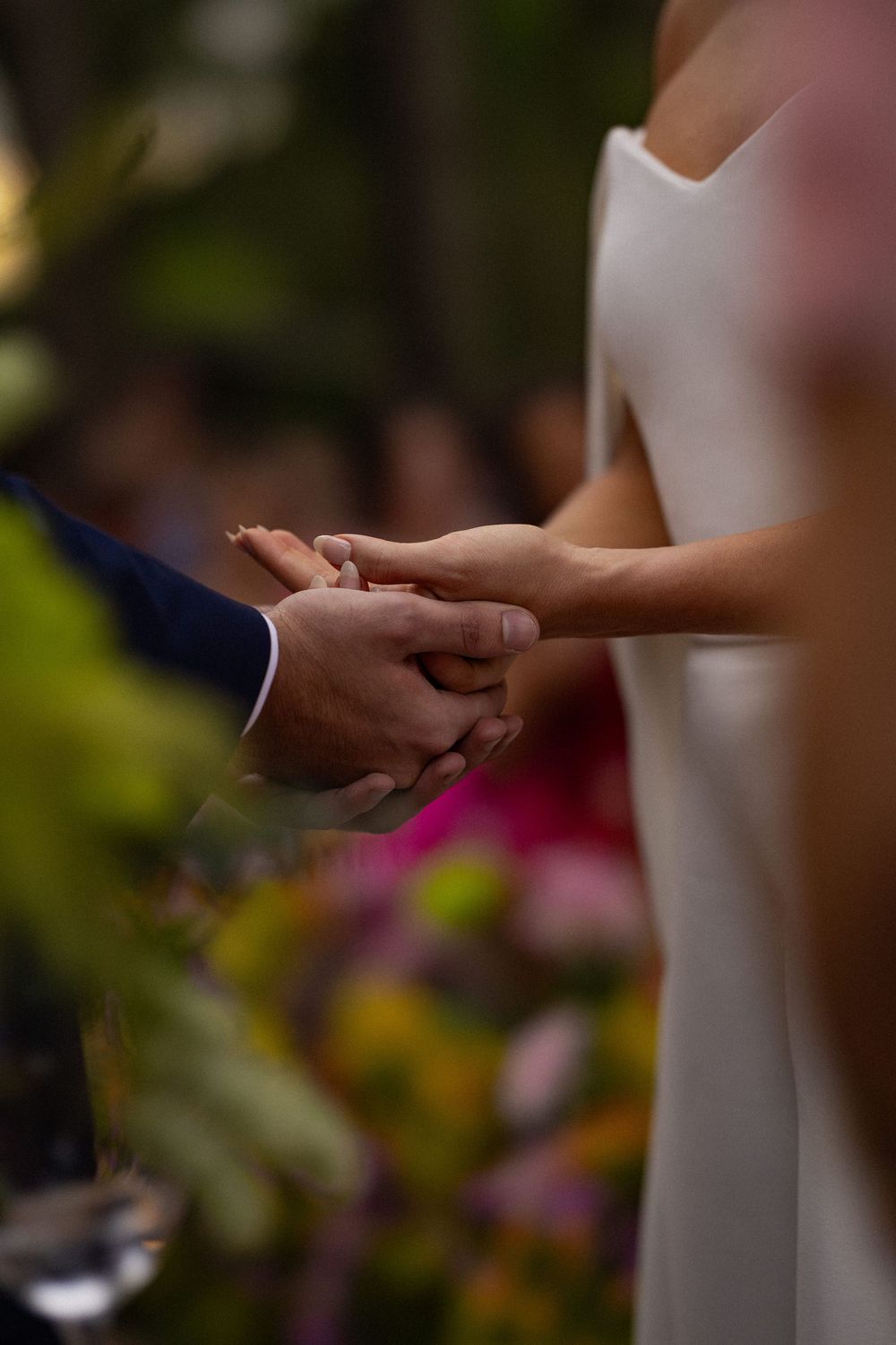 Bride in a white gown and groom in a navy suit hold hands during a wedding ceremony, with colorful flowers softly blurred behind them.