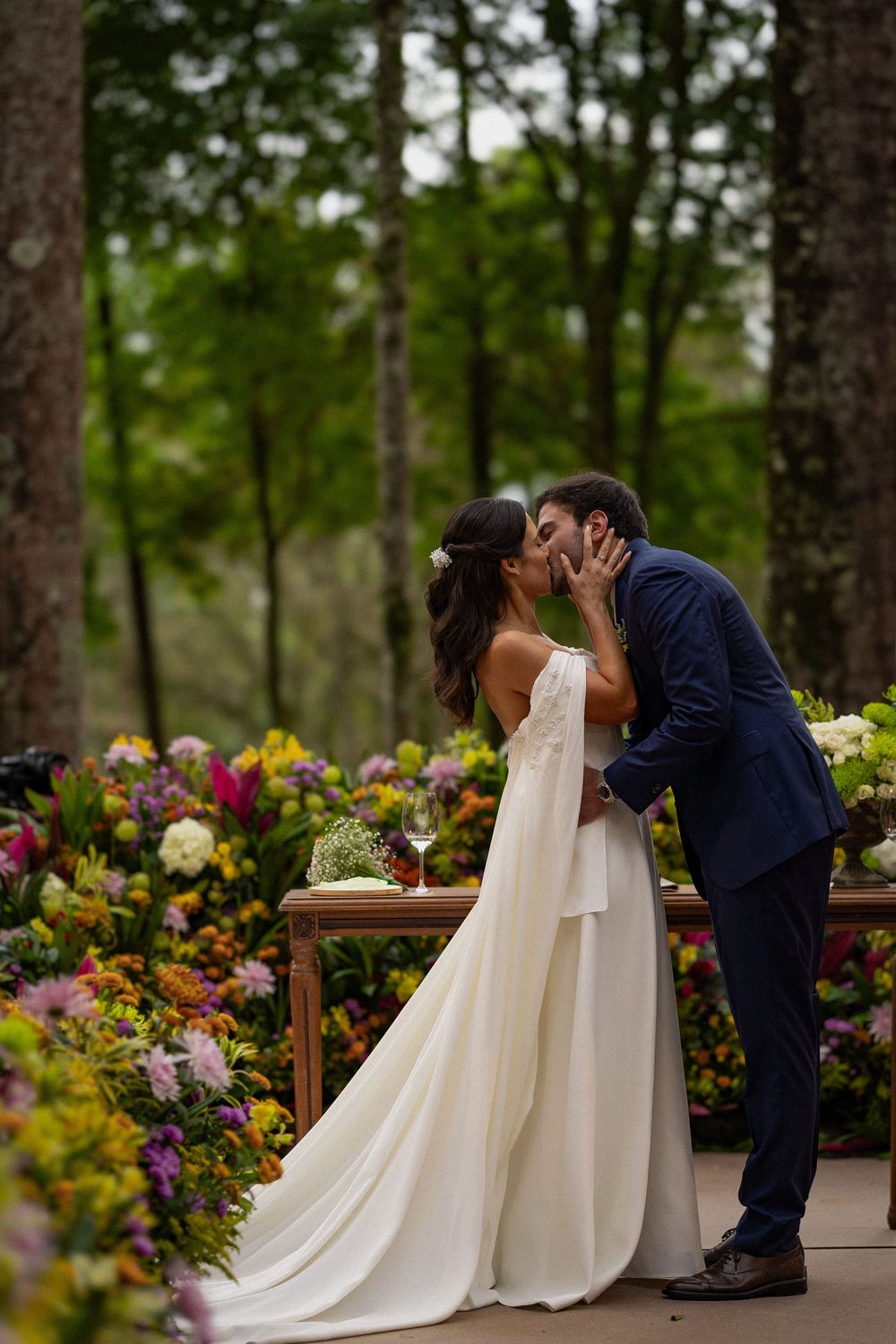 Bride in a flowing white gown and groom in a navy suit share a kiss beside a table of colorful flowers at an outdoor forest wedding.