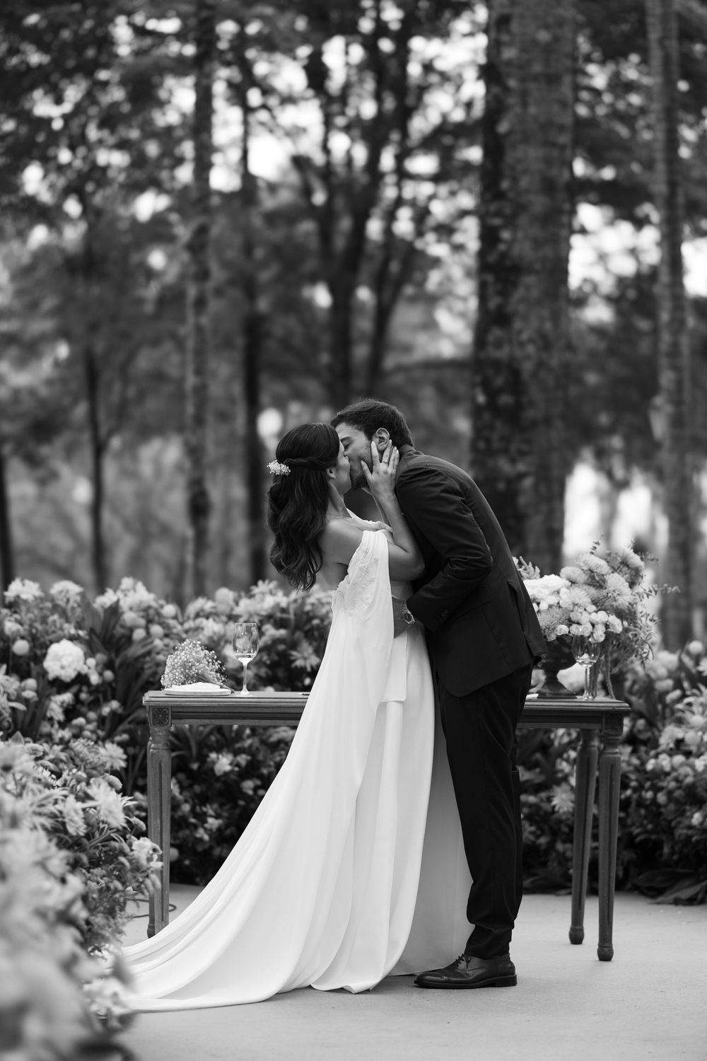 Black and white photo of a bride and groom sharing a kiss at a flower-adorned table in a wooded garden wedding ceremony.