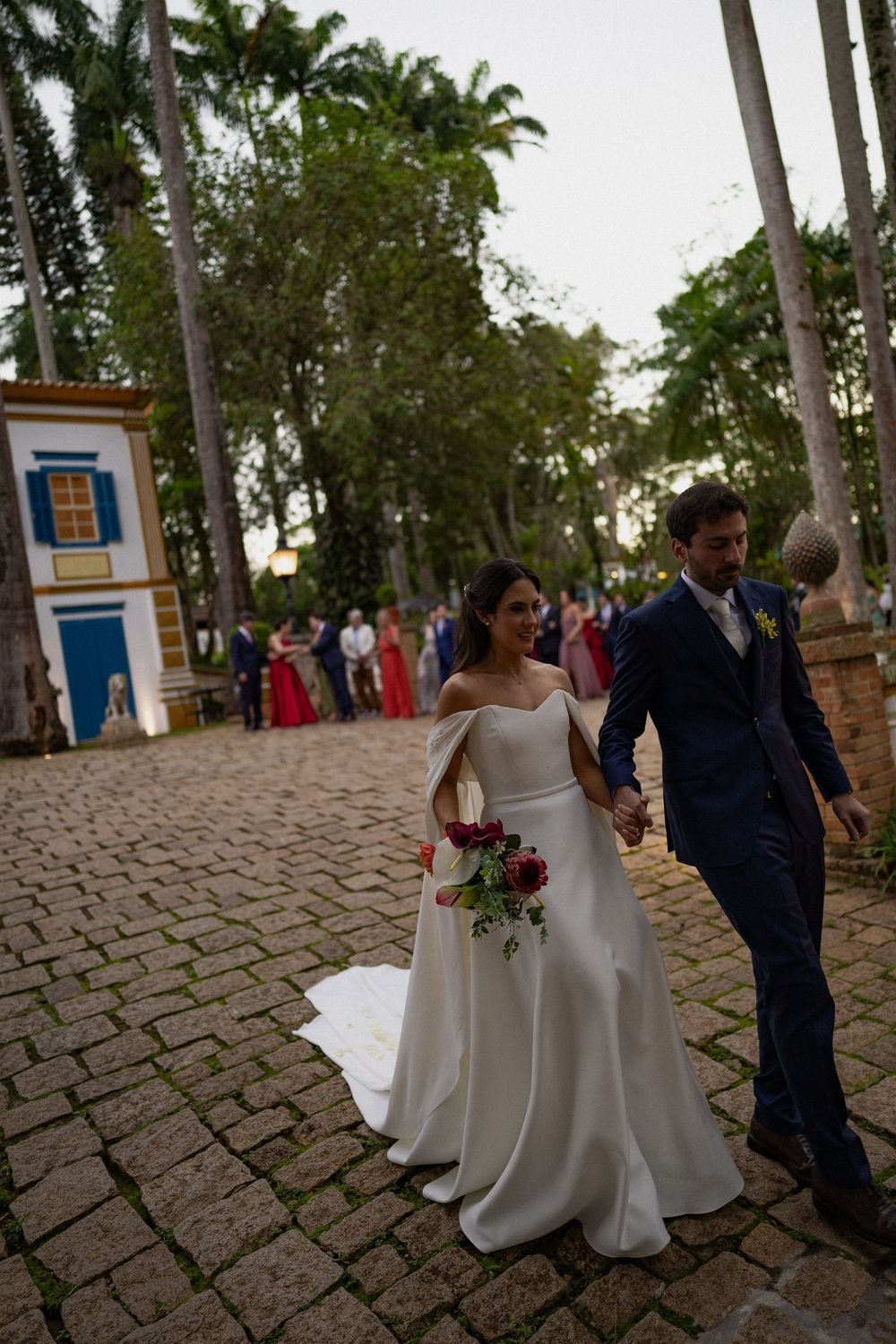 Bride in an off-shoulder white gown and groom in a navy suit hold hands on a cobblestone path as wedding guests gather under palm trees.