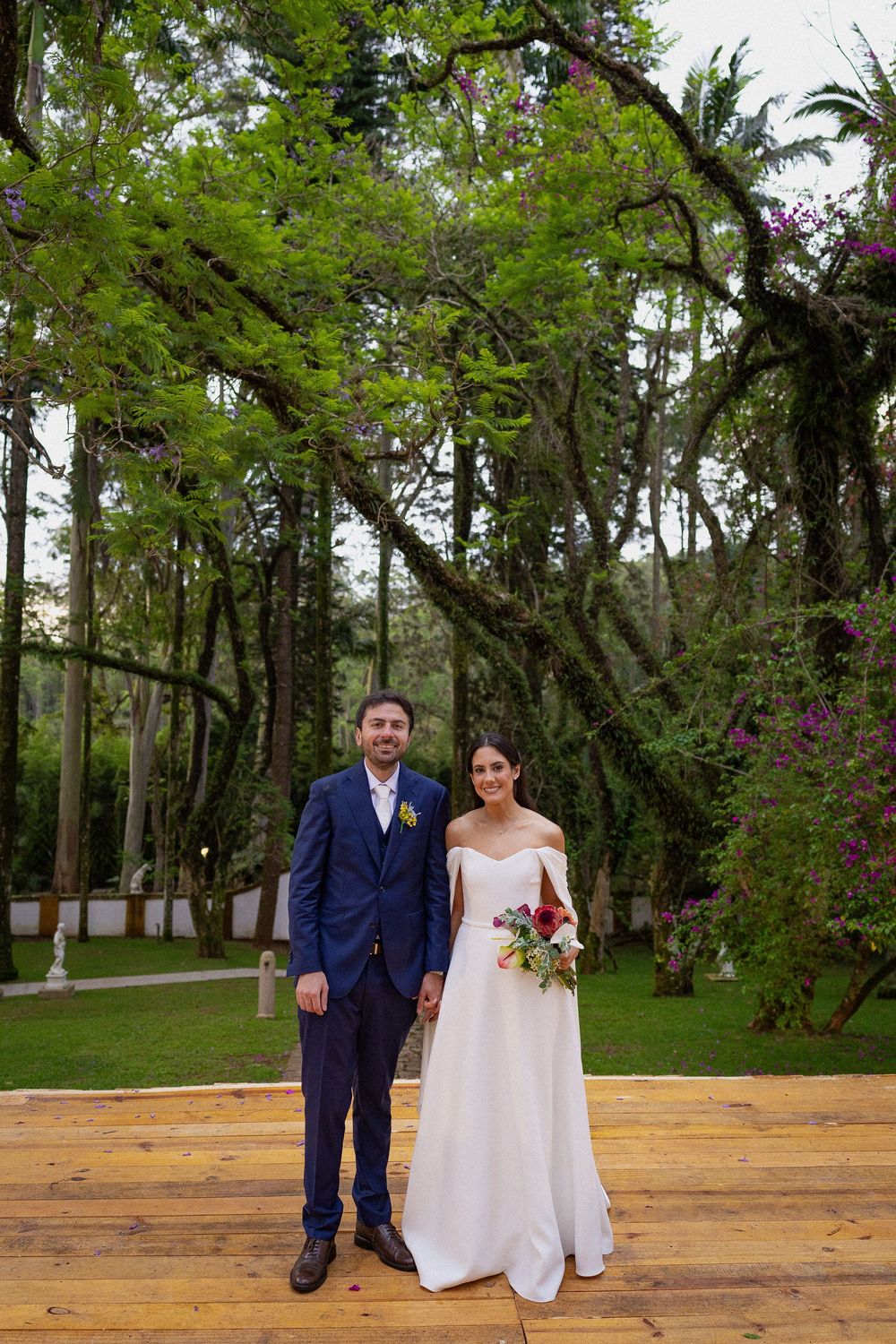Smiling bride in a white gown holding a bouquet stands beside her groom in a blue suit on a wooden deck beneath lush green trees.