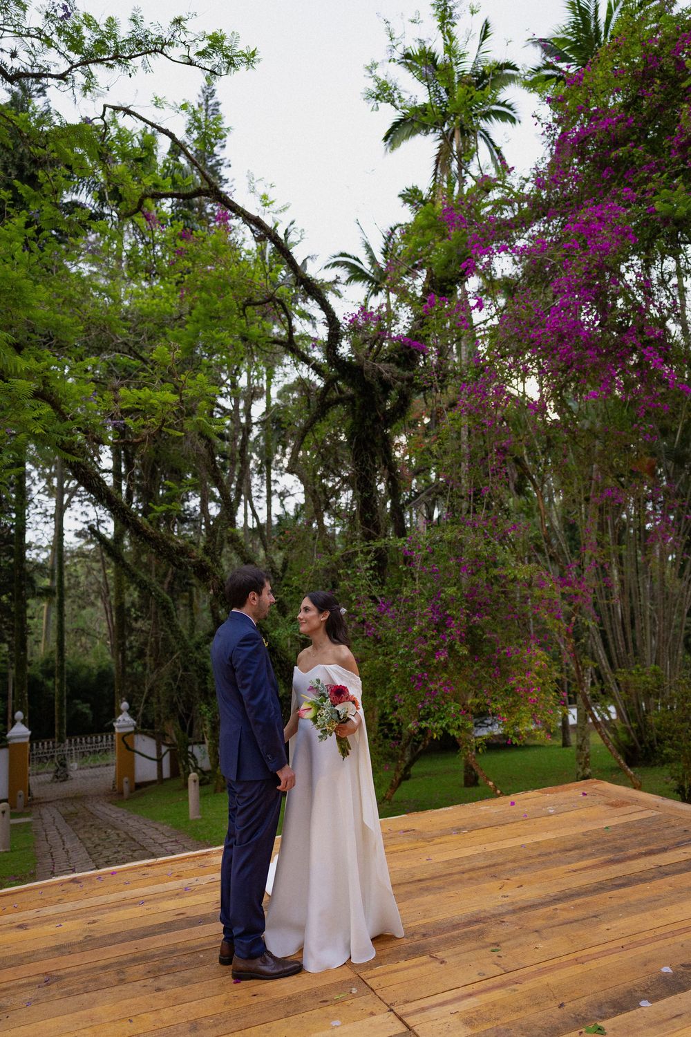 Bride in a white gown holding a bouquet faces a groom in a navy suit on a wooden deck beneath trees and purple bougainvillea.