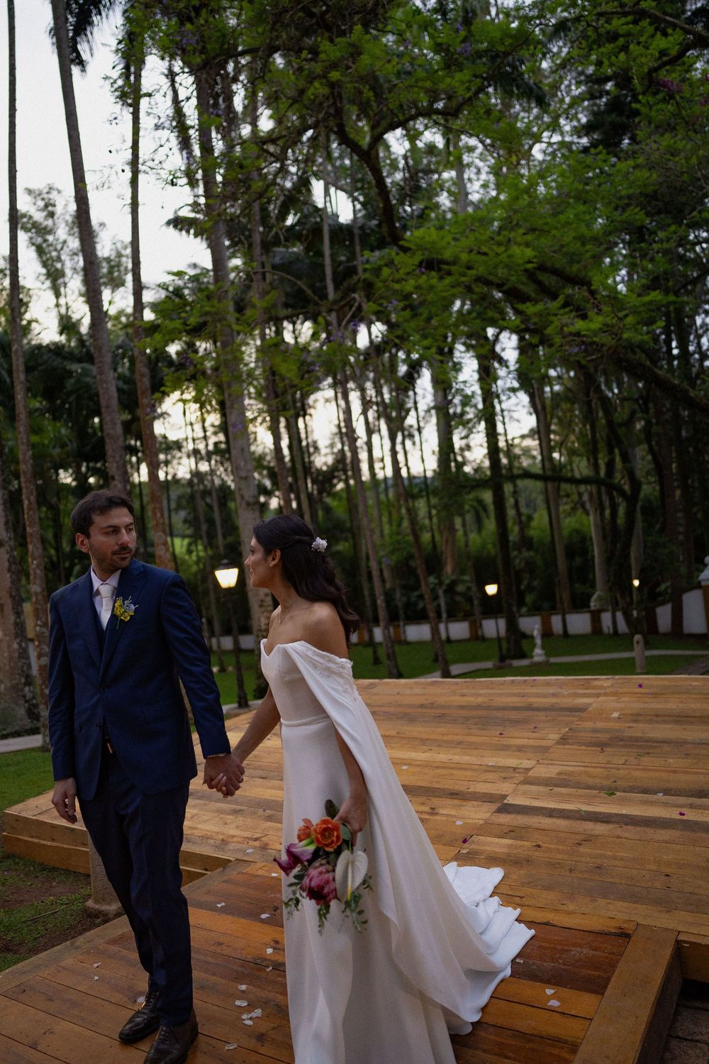 Bride in an off-shoulder white gown with cape holds hands with groom in navy suit on a wooden wedding platform among tall trees.