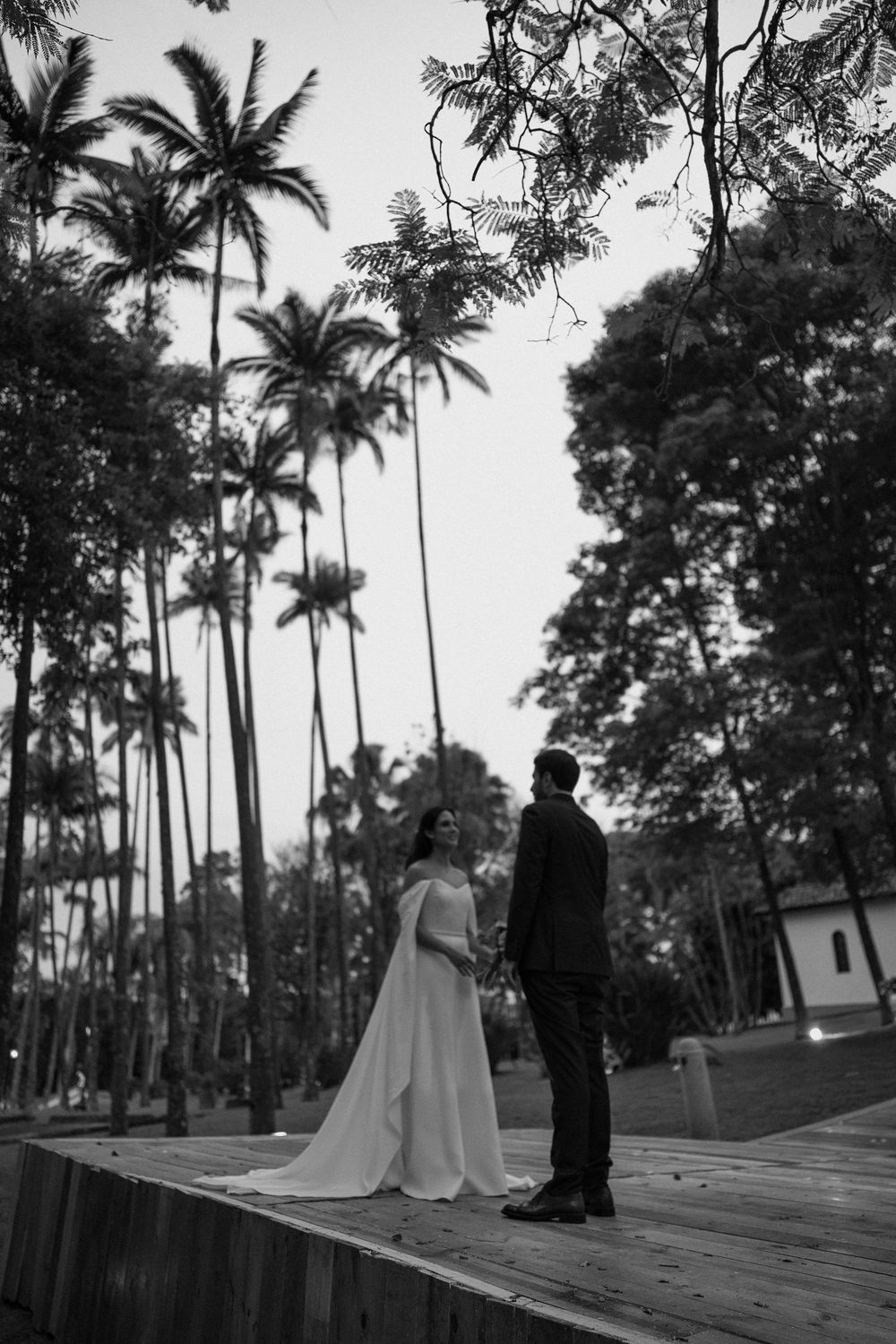 Black and white photo of a bride in a flowing gown facing a groom in a suit on a wooden platform among tall palm trees.