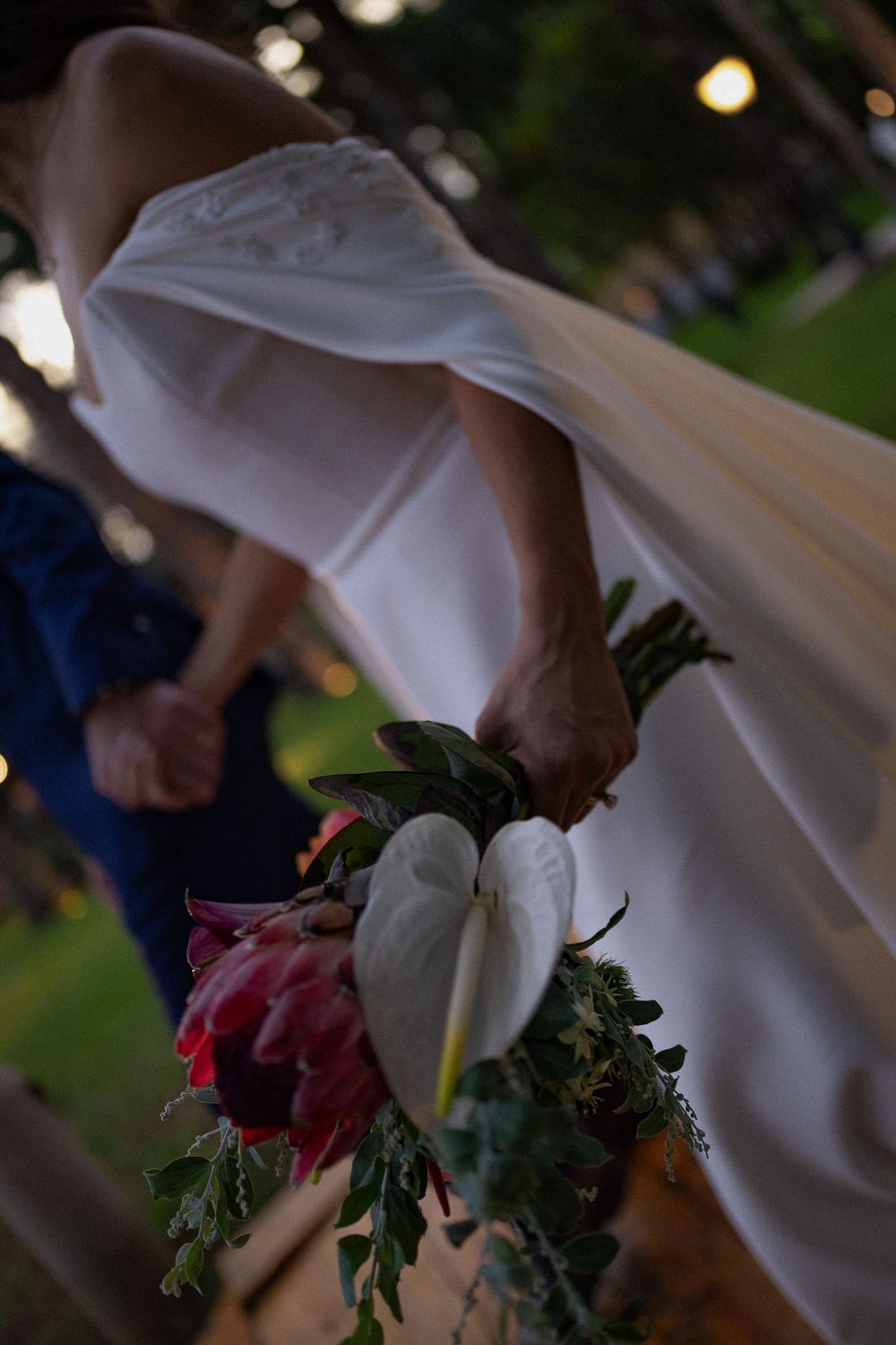Bride in a white gown holds a tropical bouquet of white anthurium, pink protea and greenery behind her back while holding hands outdoors.