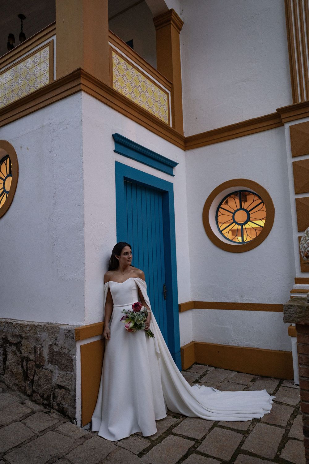 Bride in an elegant off-the-shoulder white gown with a long train holds a bouquet beside a blue door at a historic white building.
