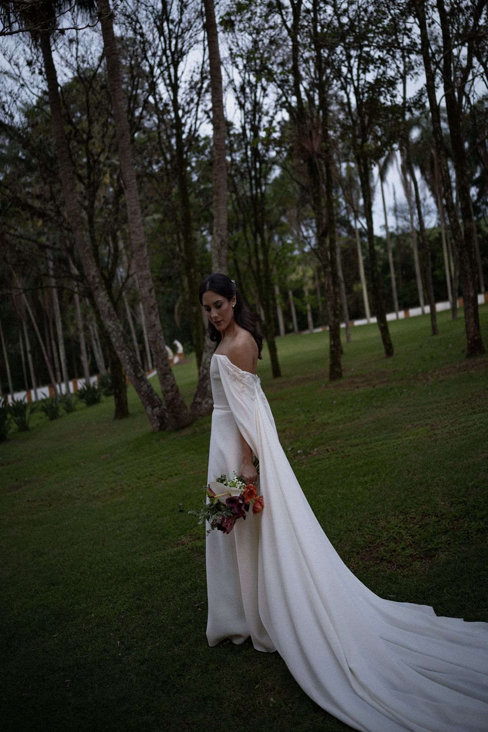 Bride in an off-the-shoulder white wedding gown with a long flowing train holds a colorful bouquet on a green lawn surrounded by tall trees.