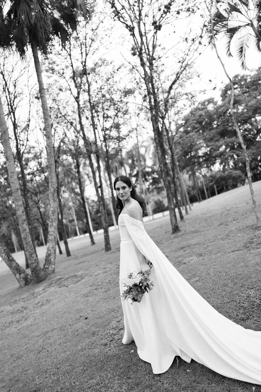 Black and white portrait of a bride in an elegant off-shoulder gown with flowing cape train, holding a bouquet in a leafy park.
