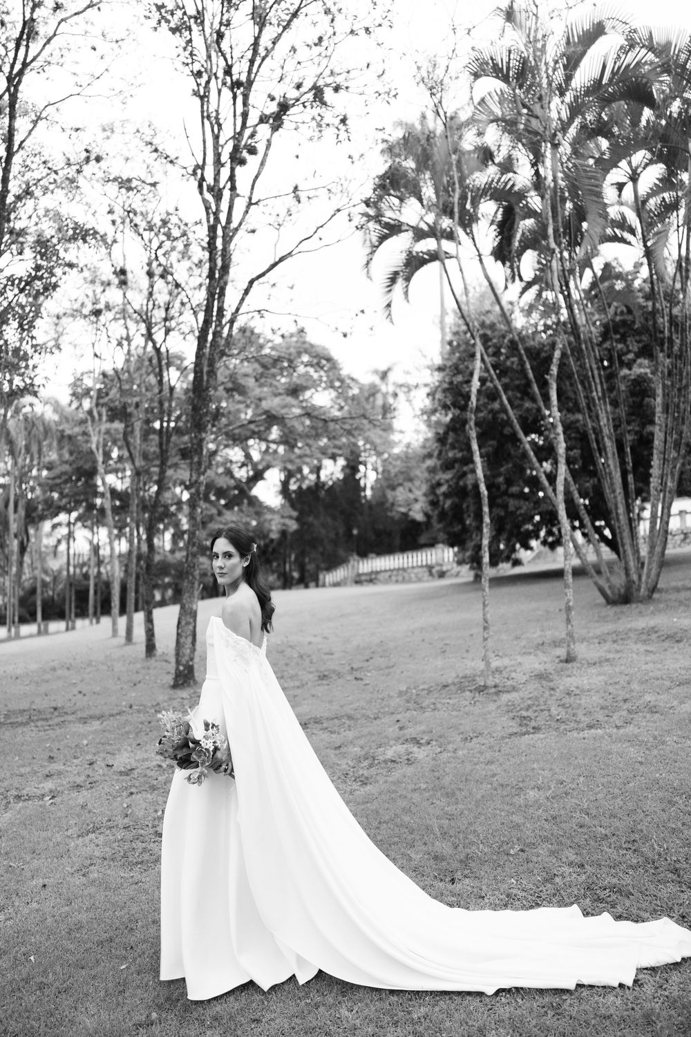 Bride in an elegant off-shoulder white gown with a long flowing train holds a bouquet while standing on a grassy lawn among tall trees.