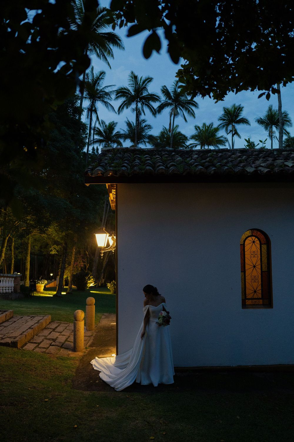 Bride in a flowing white gown stands beside a small chapel with an arched window at dusk, lit by a warm lantern among palm trees.