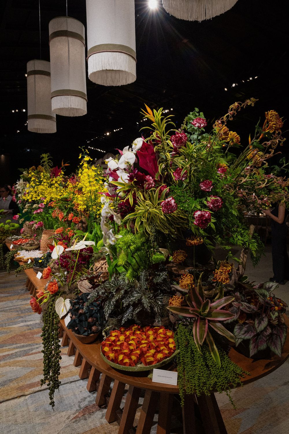 Lush floral display on a round wooden table with red, pink, and yellow blooms, tropical foliage, and hanging fringed pendant lamps.