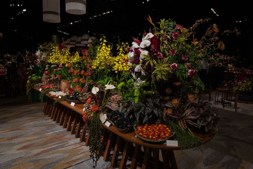 Lavish floral display on a long wooden table with yellow and white orchids, red blooms, tropical foliage and a bowl of fresh red fruit.