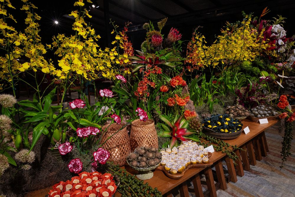 Colorful tropical flower display with yellow orchids, red anthurium, pink blooms and woven baskets of fruit on rustic wooden tables.