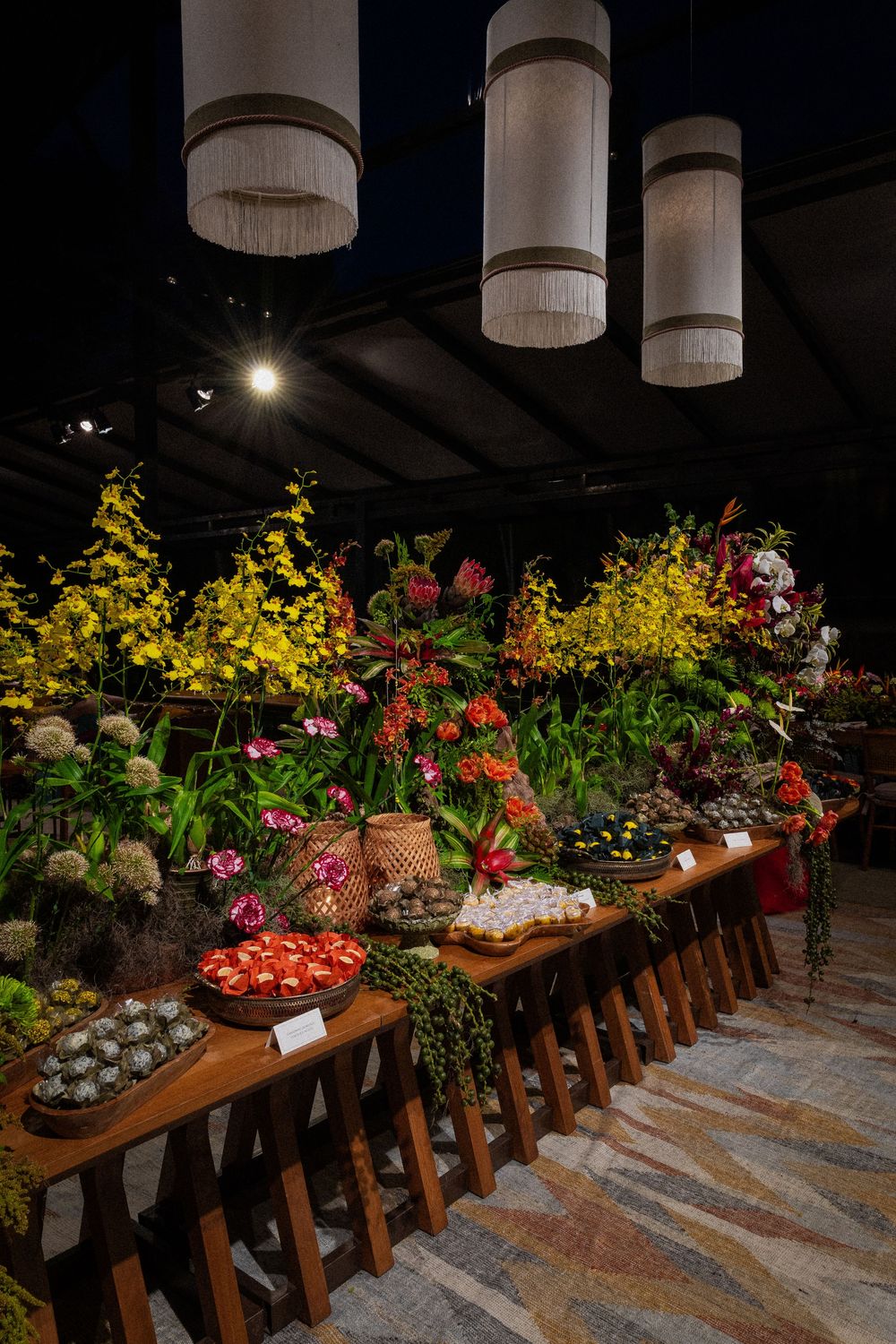 Wooden display tables hold yellow orchids, pink blooms, baskets, and fresh produce beneath modern hanging fringe pendant lights.