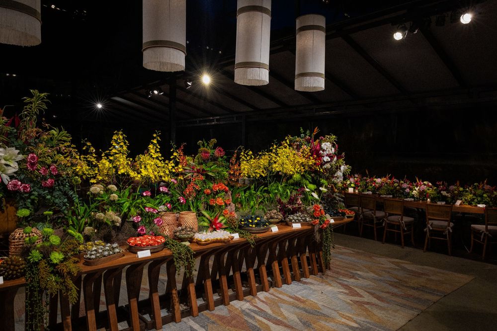 Colorful flower display of yellow, pink and red blooms arranged on long wooden tables in a dimly lit exhibition hall with hanging lanterns.