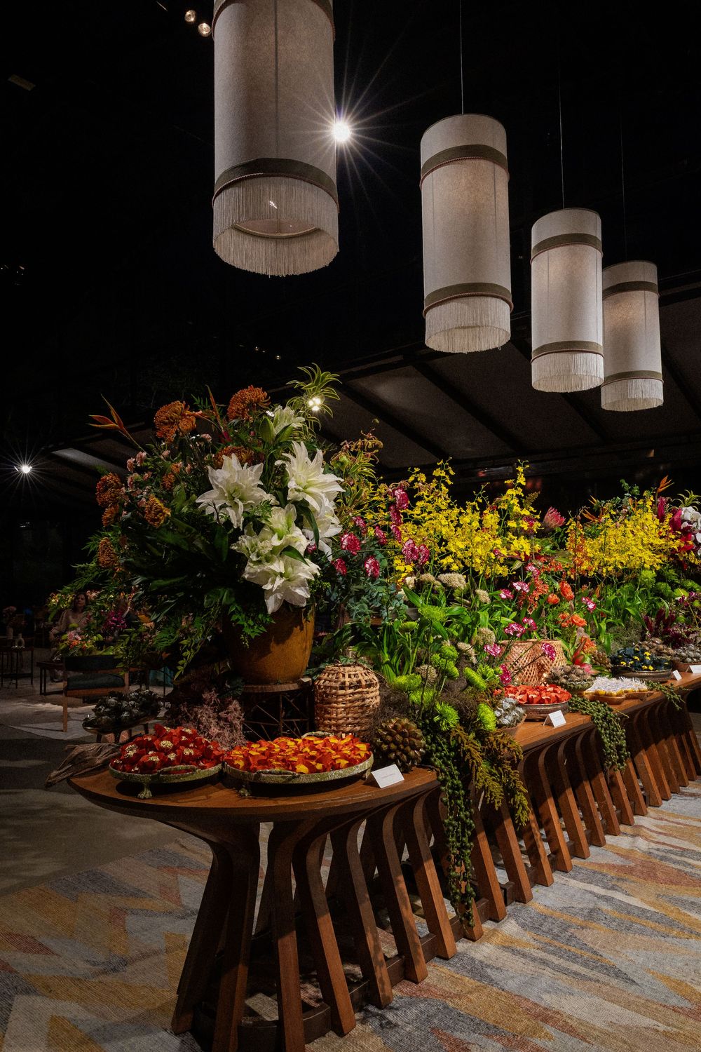 Rustic wooden table styled with lush orange, white and yellow floral arrangements and fruit platters beneath hanging fringed pendant lights.