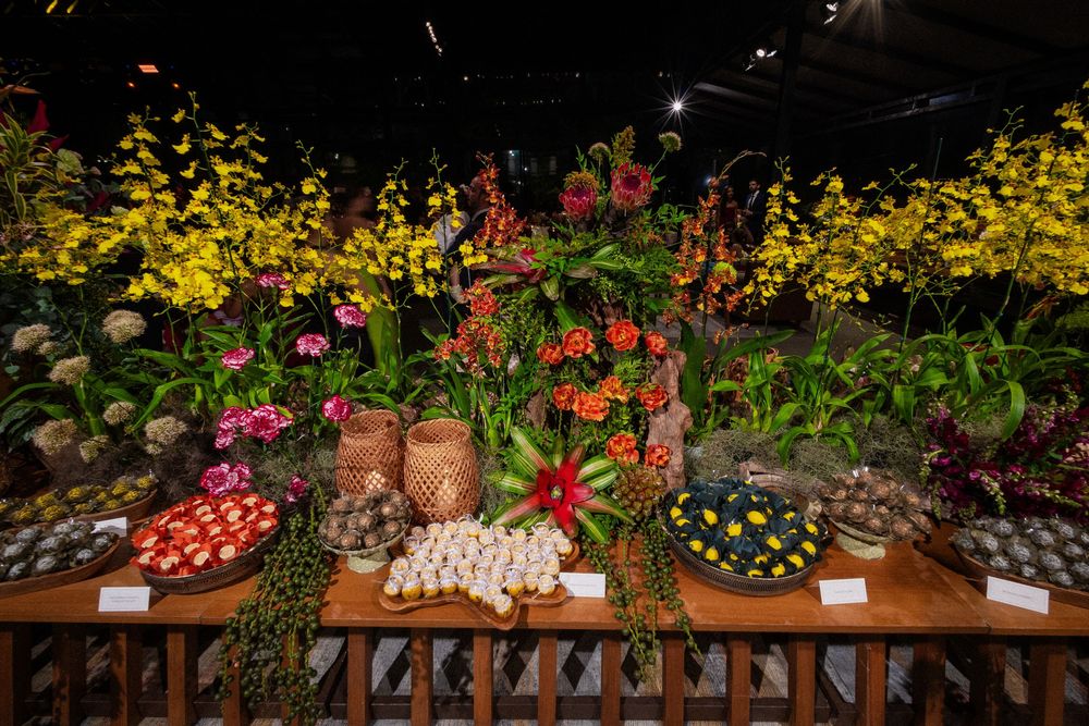 Colorful tropical flower display with yellow orchids, red anthuriums and woven baskets above a wooden table of arranged blossoms.